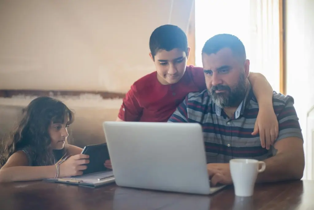 Dad using laptop with son watching on and daughter using tablet device.