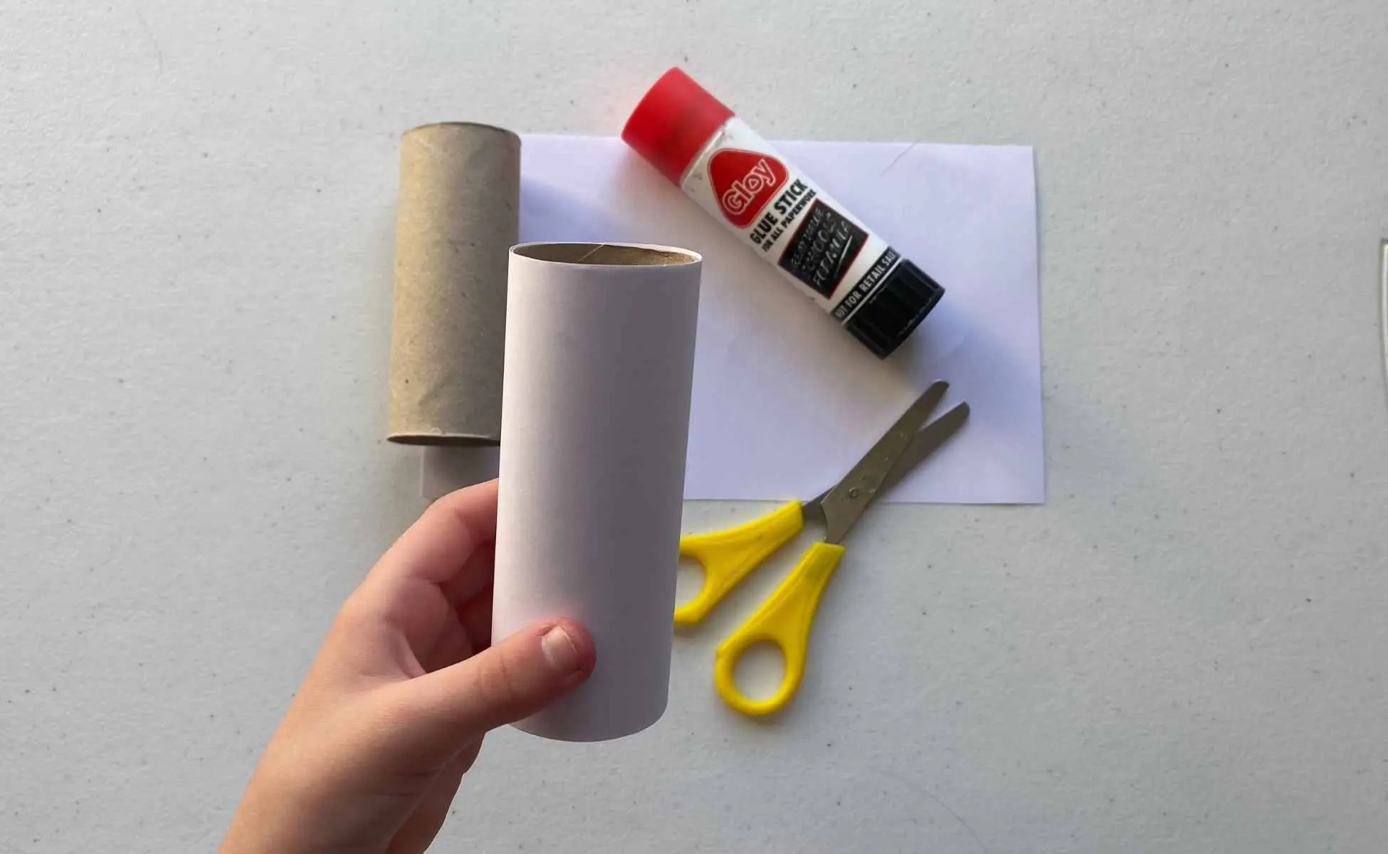 Snowman strike Christmas activity step 1 - Winston's Wish Child's hand holding toilet roll tube with glue, scissors and white paper on the table. Step one of making the snowman strike Christmas activity
