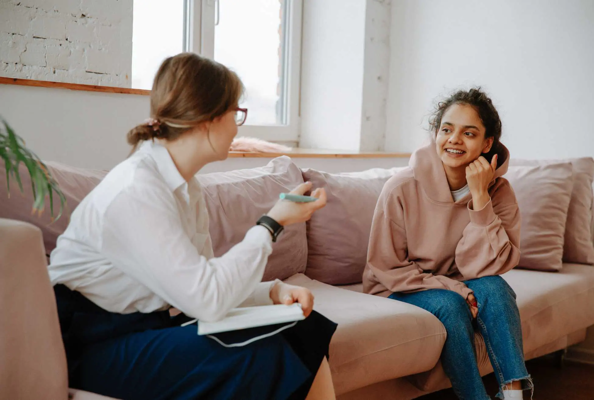 Photo of a woman holding a notebook and talking to a teenage girl sat next to her on the sofa
