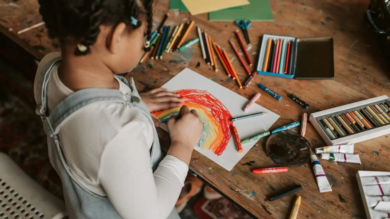 Child drawing a rainbow on tabletop with lots of art supplies around.