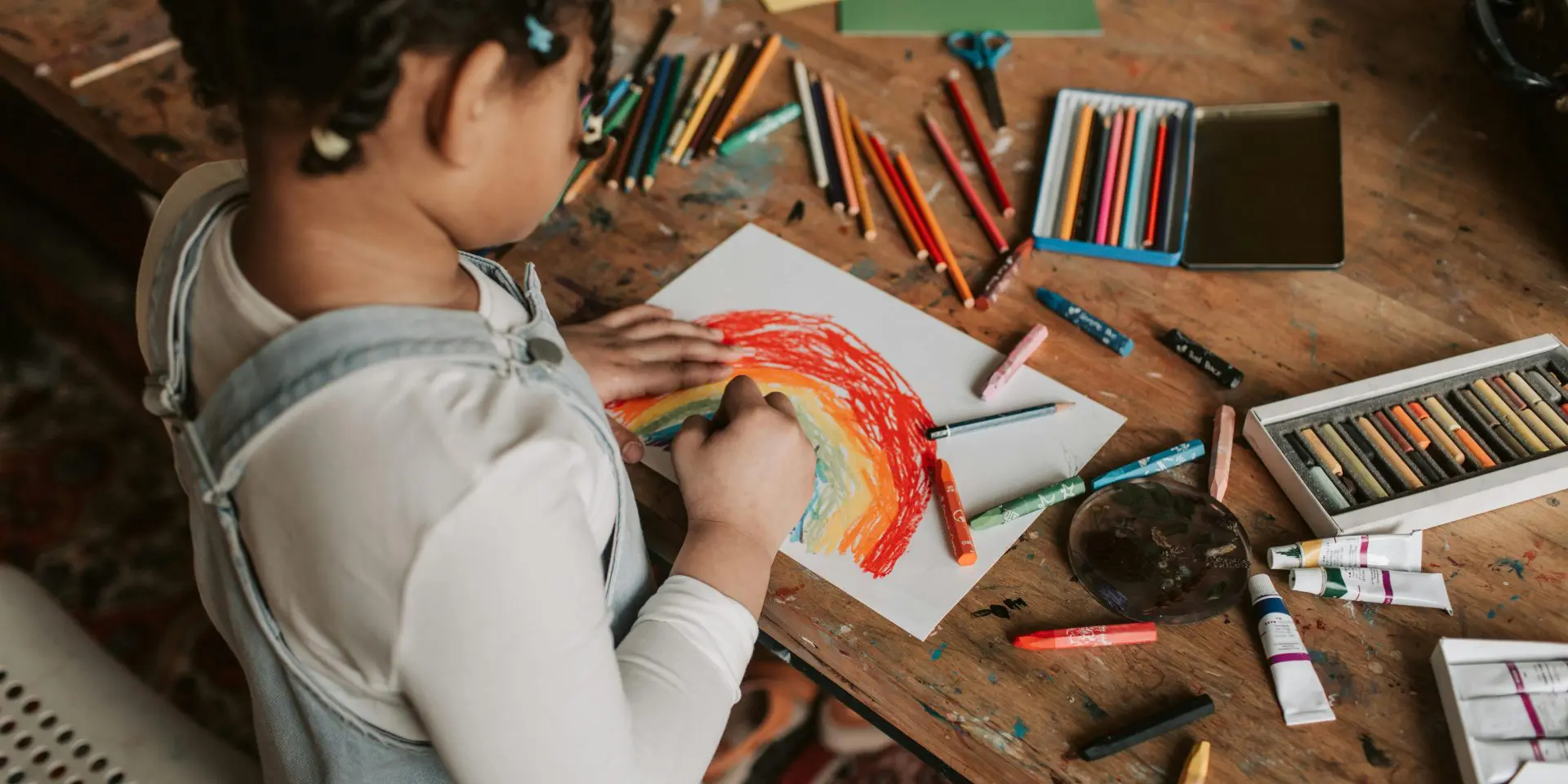 Child drawing a rainbow on tabletop with lots of art supplies around.