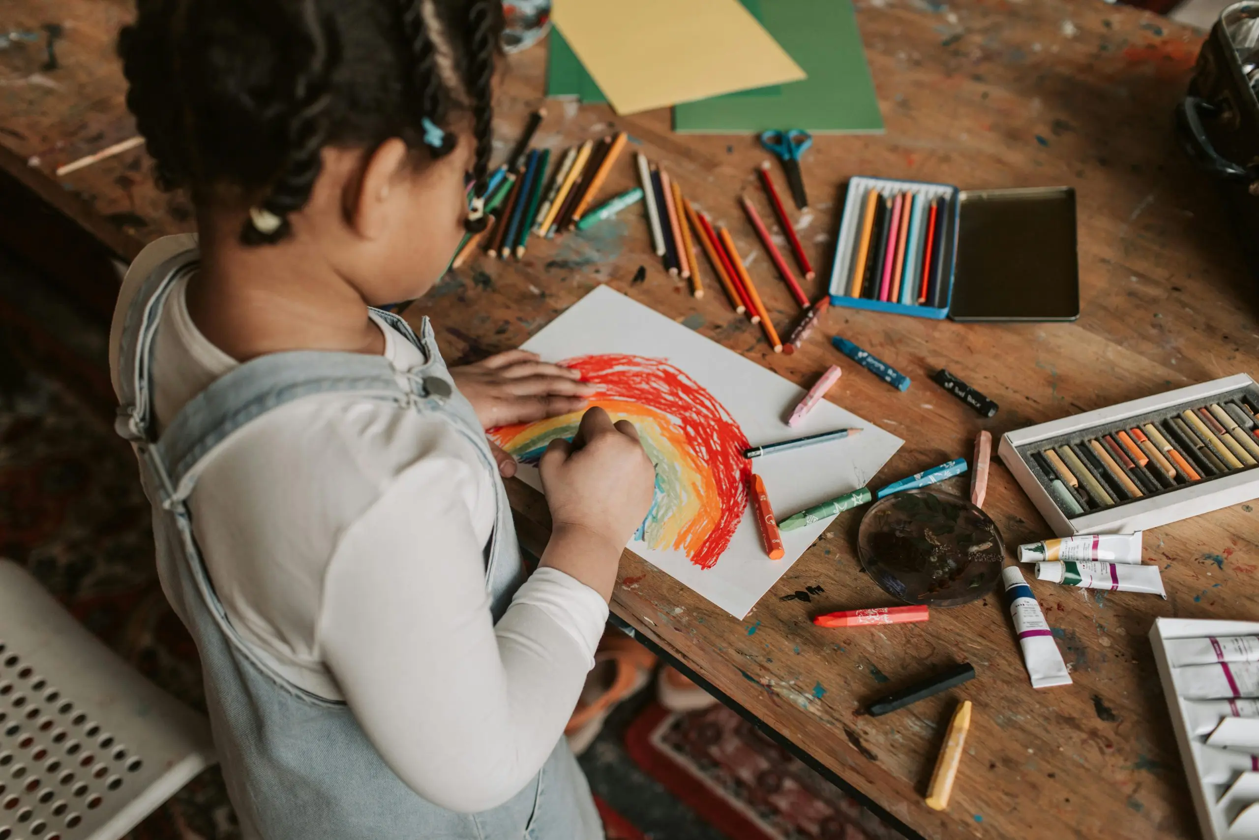 Child drawing a rainbow on tabletop with lots of art supplies around.