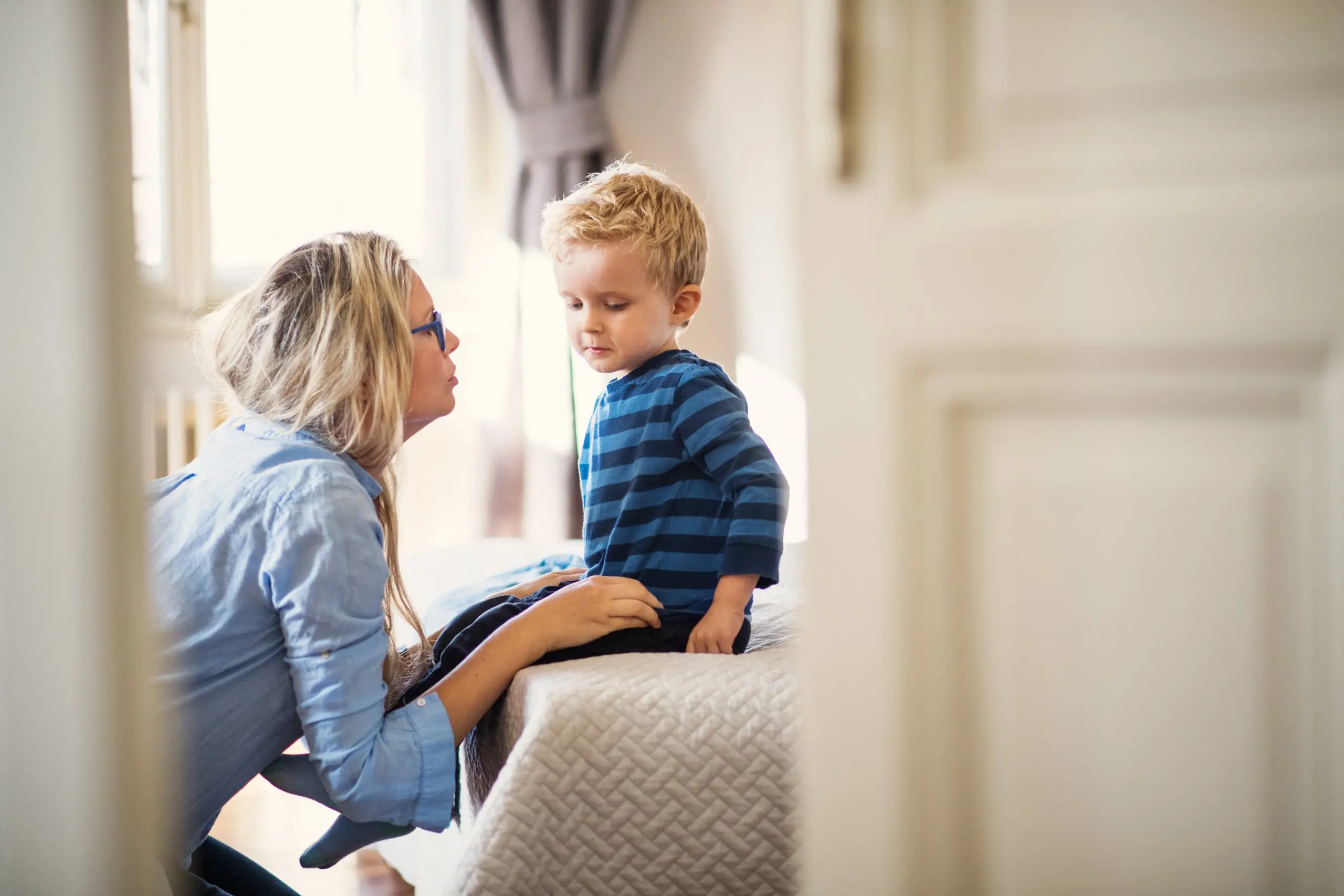 Woman kneeling talking to young boy sitting on bed.