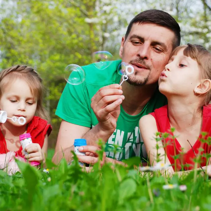 Supporting bereaved children on Mother's Day and Father's Day. Image of a man sat between his two daughters blowing bubbles on the grass