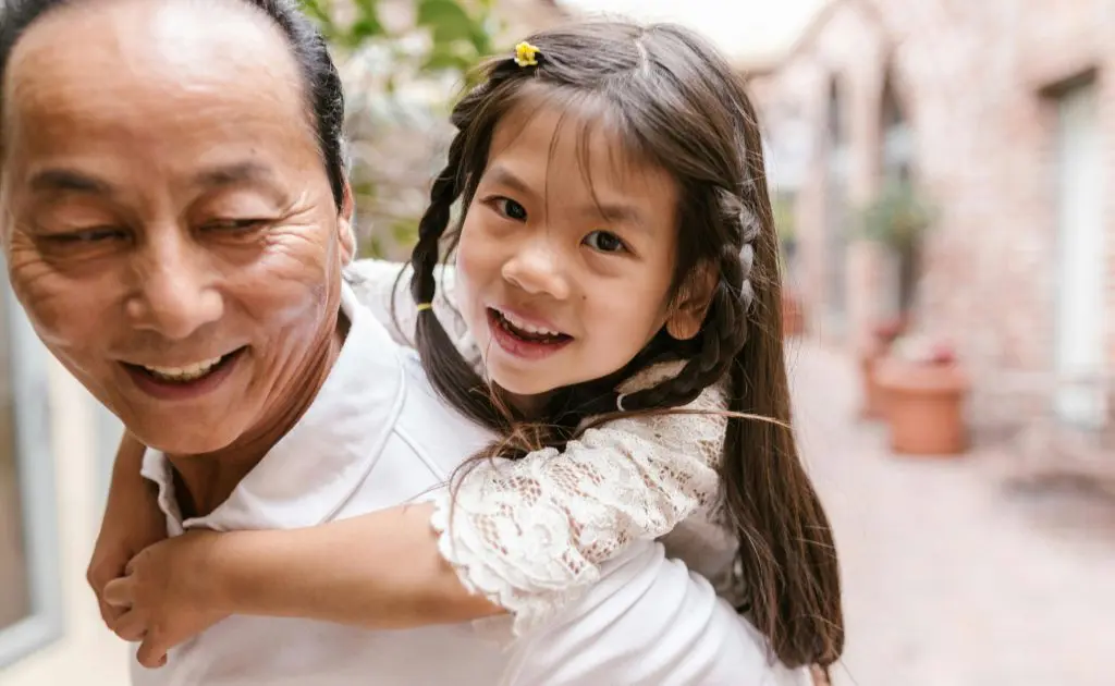 Activities for bereaved children with no memories: Image featuring an older Asian man giving a piggy back ride to a small girl with platted hair