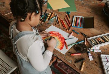 Child drawing a rainbow on tabletop with lots of art supplies around.