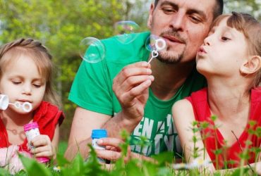 Supporting bereaved children on Mother's Day and Father's Day. Image of a man sat between his two daughters blowing bubbles on the grass